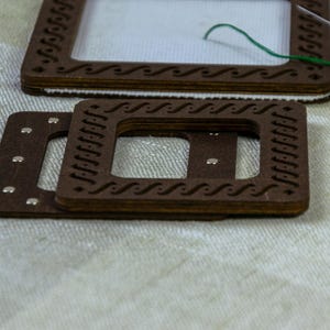 May include: Close-up of wooden embroidery hoops with decorative wave patterns. The hoops are brown and have small silver rivets. A needle with green thread is visible, suggesting a craft project. Additional wooden pieces are in the background.