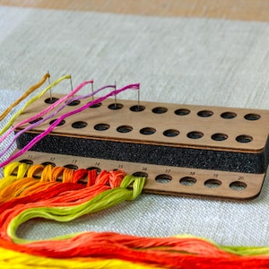 Puede incluir: Un organizador de hilo de madera con múltiples agujeros y una inserción de espuma negra. Hilo de bordar colorido en tonos de amarillo, naranja, rosa y morado está enhebrado a través del organizador, con agujas insertadas. Los suministros de manualidades están sobre una tela clara.