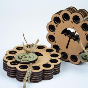 Könnte beinhalten: Zwei Fadenorganisatoren aus Holz mit blumenartigem Design. Jeder Organizer hat mehrere Löcher und besteht aus geschichtetem Holz. Einer hat eine schwarze Regenschirmgrafik. Beide haben Etiketten mit Bindfäden befestigt.