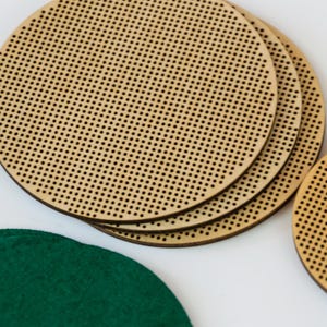 May include: Several round, wooden cross-stitch hoops with small, evenly spaced holes. A few are stacked together, and a green felt circle is visible in the lower left corner. The hoops are made of light-colored wood.