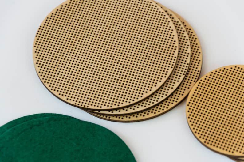 May include: Several round, wooden cross-stitch hoops with small, evenly spaced holes. A few are stacked together, and a green felt circle is visible in the lower left corner. The hoops are made of light-colored wood.