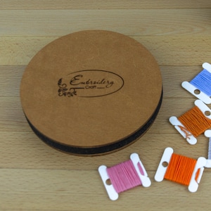 May include: A round, brown embroidery craft case with the text "Embroidery Craft" and several spools of colorful thread. The spools are white with blue, orange, pink, and gray thread. The case is on a wooden surface.