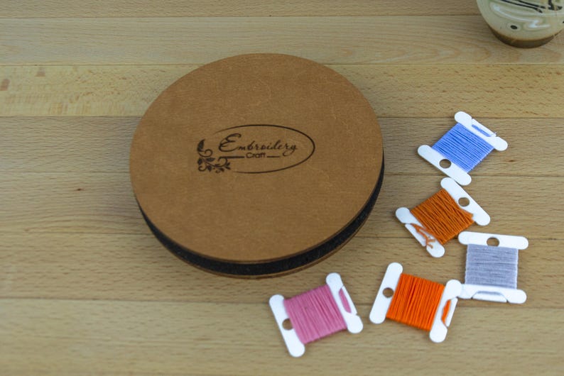 May include: A round, brown embroidery craft case with the text "Embroidery Craft" and several spools of colorful thread. The spools are white with blue, orange, pink, and gray thread. The case is on a wooden surface.