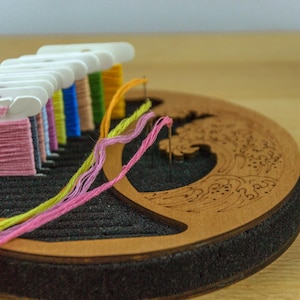 May include: A wooden embroidery floss organizer with a wave design, holding spools of colorful thread. Pink, yellow, blue, and green threads are visible, with needles inserted. The organizer sits on a wooden surface, ideal for craft projects.