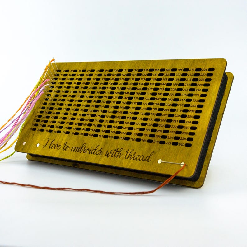 May include: A wooden embroidery floss organizer with a grid of numbered holes. The organizer is stained a golden color and has the phrase "I love to embroider with thread" etched on the front. Colorful threads are attached.