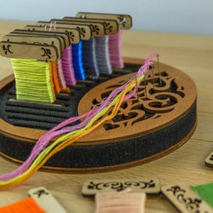 Pode incluir: Um organizador de linha de bordar de madeira com um design yin-yang. O organizador contém carretéis de linha de bordar colorida em tons de verde, laranja, rosa, azul e cinza. A linha é disposta em pequenos carretéis de madeira, com alguns fios se estendendo.