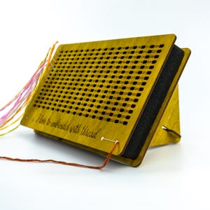 May include: A wooden embroidery floss organizer with a grid of numbered holes. The organizer is angled, with a black foam insert and the text "I love to embroider with thread". Colorful embroidery floss is attached.