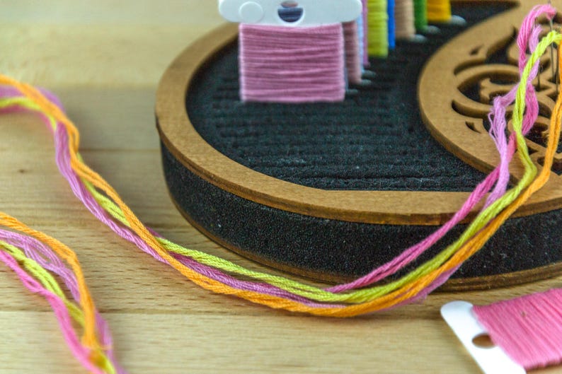 May include: Close-up of colorful embroidery floss in pink, yellow, and orange. The floss is next to a wooden thread organizer with a black interior and a brown wooden rim. The background is a light wooden surface.
