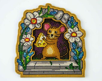 May include: A cross-stitch embroidery piece on a wooden frame featuring a brown mouse holding a yellow piece of Swiss cheese. The mouse is framed by a stone archway decorated with white flowers, green leaves, and blue blossoms.