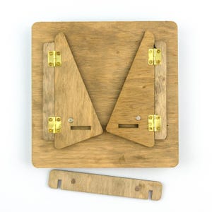Op de afbeelding: Een houten ezel met twee driehoekige panelen die plat inklappen. De ezel heeft aan elke kant twee messing scharnieren en een kleine houten balk die de twee panelen verbindt.