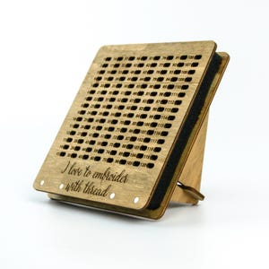 Op de afbeelding: Houten draadorganizer met een zwarte schuimrug en een standaard. De organizer heeft een raster met genummerde gaten voor het opbergen van borduurgaren. De tekst "Ik hou van borduren met draad" is gegraveerd op de voorkant.