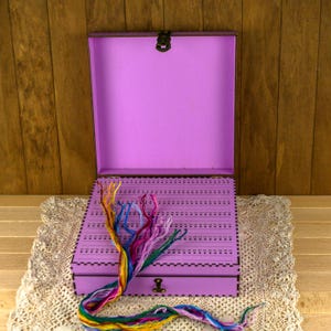Può includere: Scatola di legno viola con un cassetto e un interno a griglia per riporre il filo da ricamo. La scatola è aperta e un arcobaleno di filo ne fuoriesce.