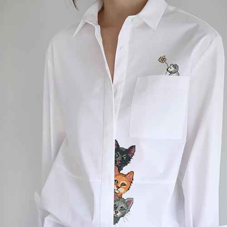 May include: White long-sleeve button-down shirt with a pocket on the left side. The pocket has an embroidered image of a mouse holding a flower. The shirt has an embroidered image of three cats peeking out from behind the button placket.