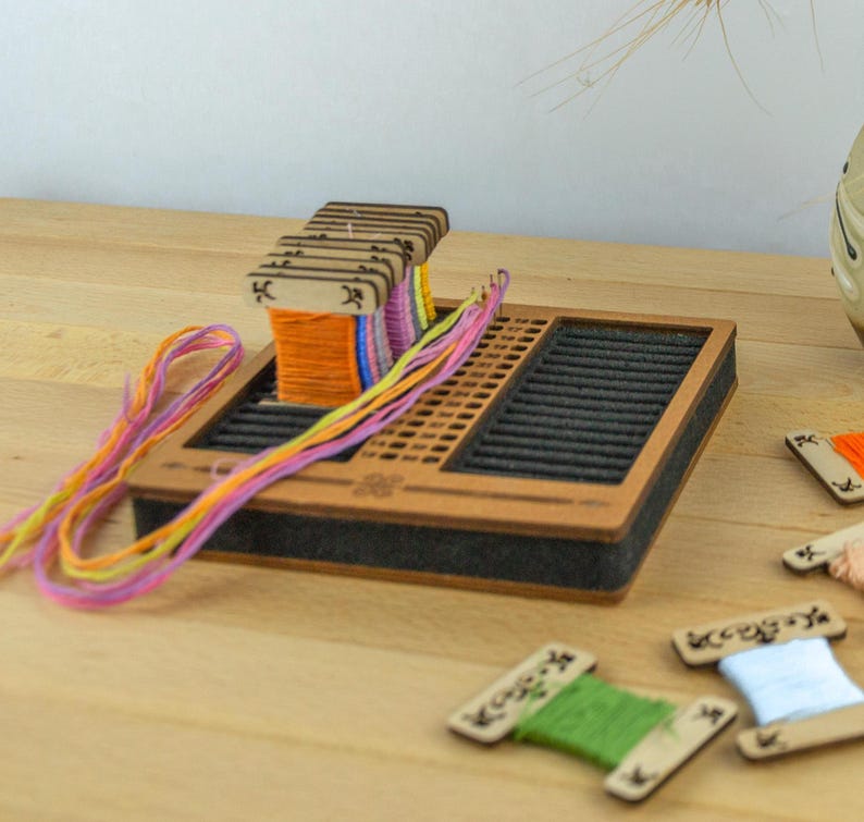 May include: A wooden embroidery floss organizer with a brown frame and black interior. The organizer holds spools of colorful embroidery floss. Several wooden bobbins with green, orange, and blue floss are scattered around the organizer.