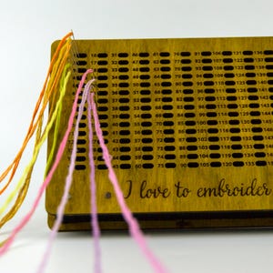 May include: Wooden embroidery tool with numbered holes and colorful threads. The text "I love to embroider" is visible on the wooden surface. Needles are inserted through the holes, with threads in shades of pink, yellow, and orange.