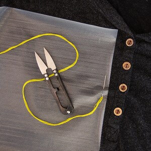 May include: A close-up of a yellow thread and a pair of small scissors on a white mesh fabric. The fabric is next to a black shirt with brown buttons.