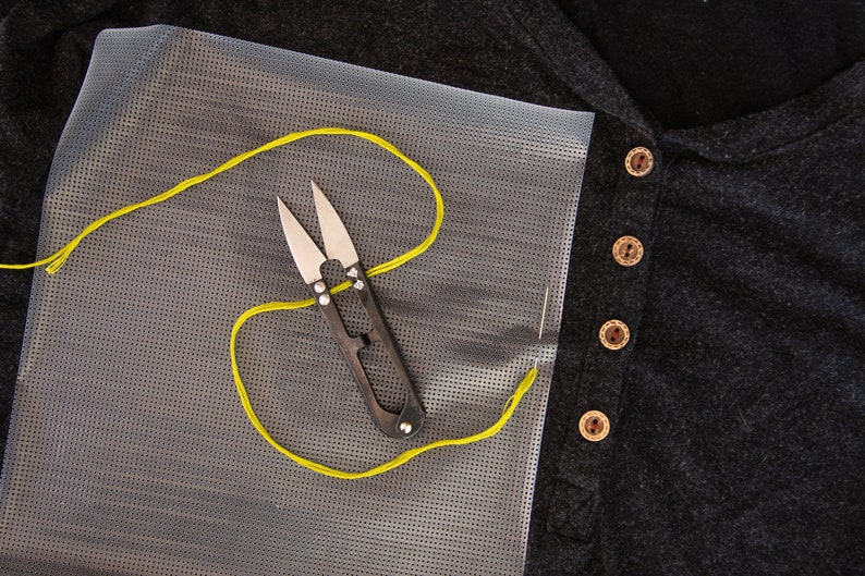 May include: A close-up of a yellow thread and a pair of small scissors on a white mesh fabric. The fabric is next to a black shirt with brown buttons.