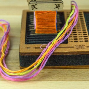 May include: A wooden friendship bracelet maker with a spool of orange thread and colorful strands of yellow, pink, and purple thread. The wooden base has a black foam edge and decorative etched designs.