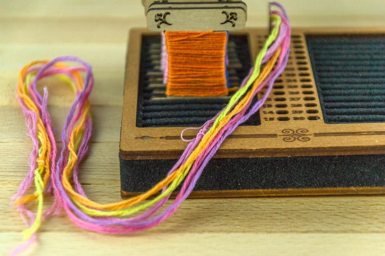 May include: A wooden friendship bracelet maker with a spool of orange thread and colorful strands of yellow, pink, and purple thread. The wooden base has a black foam edge and decorative etched designs.