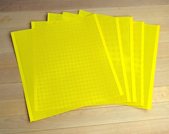 May include: A set of five square sheets of bright yellow plastic canvas, each featuring a uniform grid of small, evenly spaced holes throughout the entire surface, arranged in a slightly fanned, overlapping stack on a flat surface.