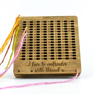 Op de afbeelding: Een houten borduurdraadorganizer met een genummerd rooster om tot 120 spoelen draad op te bergen. De organizer heeft roze, oranje, gele en bruine draad eromheen gewikkeld. De tekst "Ik hou van borduren met draad" is gegraveerd op de voorkant van de organizer.