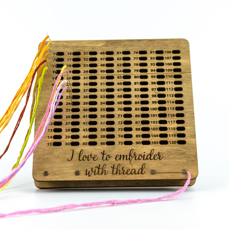 Op de afbeelding: Een houten borduurdraadorganizer met een genummerd rooster om tot 120 spoelen draad op te bergen. De organizer heeft roze, oranje, gele en bruine draad eromheen gewikkeld. De tekst "Ik hou van borduren met draad" is gegraveerd op de voorkant van de organizer.