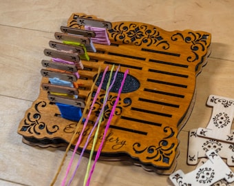 May include: A wooden embroidery floss organiser with laser-cut floral patterns and the word Embroidery engraved on the surface. It features multiple slots holding colourful thread bobbins with pink, yellow, blue, and orange floss, plus extra bobbins.