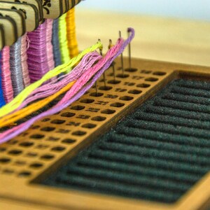 May include: A wooden embroidery floss organizer with multiple colors of thread. The organizer has a series of holes for holding the floss and a black felt area. The floss colors include orange, blue, purple, yellow, and pink.
