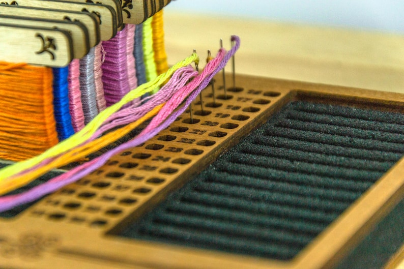 May include: A wooden embroidery floss organizer with multiple colors of thread. The organizer has a series of holes for holding the floss and a black felt area. The floss colors include orange, blue, purple, yellow, and pink.