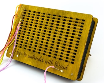 May include: A rectangular yellow wooden embroidery thread organiser featuring a grid of 180 numbered holes. The text "I love to embroider with thread" is engraved on the front. Several strands of colourful embroidery floss are threaded through the holes.