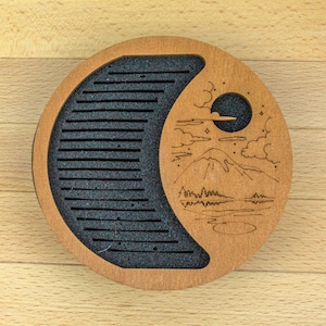 May include: A circular wooden tea tray with a crescent-shaped section filled with black foam and horizontal grooves. The other half features an etched mountain and moon design. The tray is light brown.