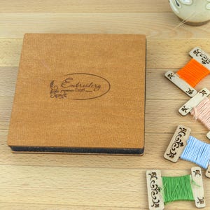 May include: A wooden box with the word "Embroidery Craft" and decorative spools of thread in orange, pink, blue, and green. The spools are on a light wooden surface.