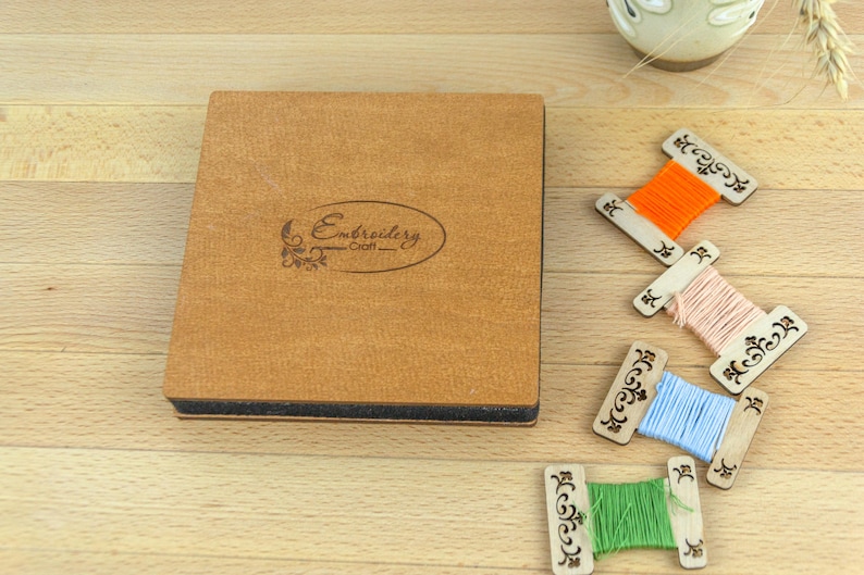 May include: A wooden box with the word "Embroidery Craft" and decorative spools of thread in orange, pink, blue, and green. The spools are on a light wooden surface.