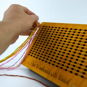 May include: A wooden embroidery board with a grid of numbered holes and a message that reads "I love to embroider with thread".  The board is being used to thread colourful embroidery floss.