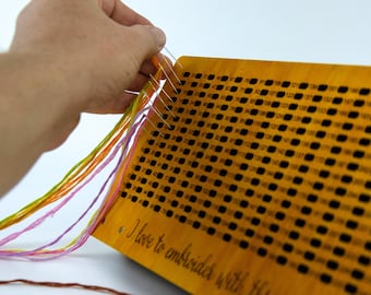 May include: A wooden embroidery board with a grid of numbered holes and a message that reads "I love to embroider with thread".  The board is being used to thread colorful embroidery floss.