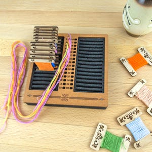 May include: Wooden embroidery floss organizer with slots and a stack of thread cards. Colorful embroidery floss in pink, yellow, and orange is visible, along with several spools of thread in various colors. A decorative vase is in the background.