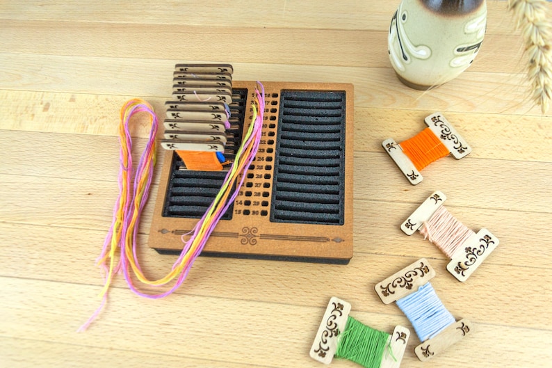 May include: Wooden embroidery floss organizer with slots and a stack of thread cards. Colorful embroidery floss in pink, yellow, and orange is visible, along with several spools of thread in various colors. A decorative vase is in the background.