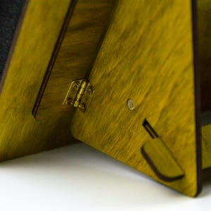 May include: Close-up of a wooden, triangular-shaped object with a yellow-green stain. The object is connected by a brass hinge. A small, circular magnet and a rectangular cutout are visible on the side.