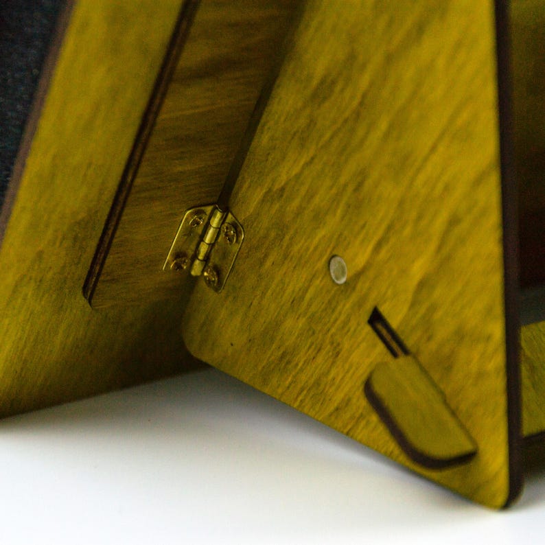 May include: Close-up of a wooden, triangular-shaped object with a yellow-green stain. The object is connected by a brass hinge. A small, circular magnet and a rectangular cutout are visible on the side.