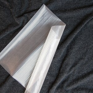 May include: A white plastic sheet with a grid pattern. The sheet is folded in half and lying on a dark gray fabric surface.