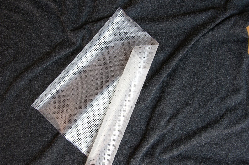 May include: A white plastic sheet with a grid pattern. The sheet is folded in half and lying on a dark gray fabric surface.