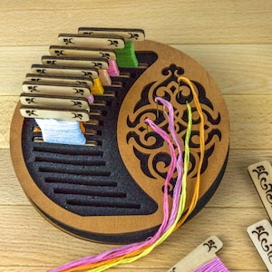 Op de afbeelding: Een houten draadorganizer met een decoratief uitgesneden ontwerp. De organizer bevat spoelen met kleurrijk garen, met strengen roze, gele en oranje garen die zich vanuit het midden uitstrekken. Extra garenspoelen liggen in de buurt verspreid.