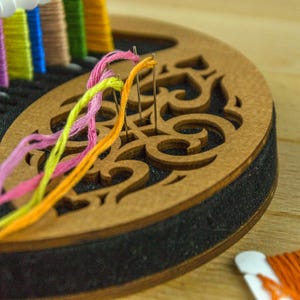 May include: A wooden embroidery floss organizer with a decorative cutout design. Colorful embroidery floss in pink, yellow, orange, and green threads are threaded through needles. Other spools of floss are visible.