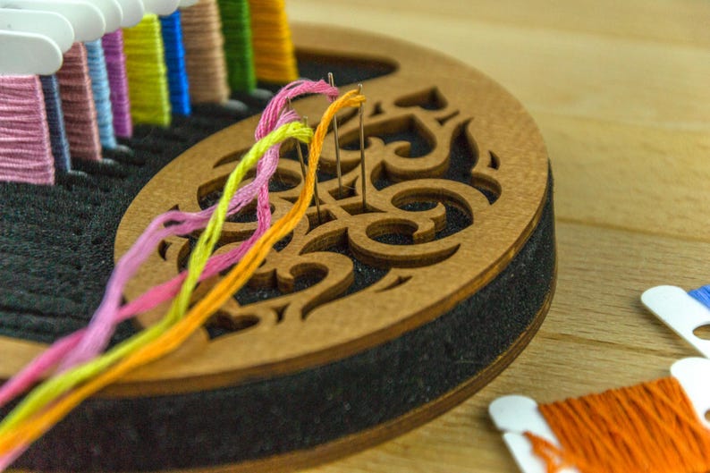 May include: A wooden embroidery floss organizer with a decorative cutout design. Colorful embroidery floss in pink, yellow, orange, and green threads are threaded through needles. Other spools of floss are visible.