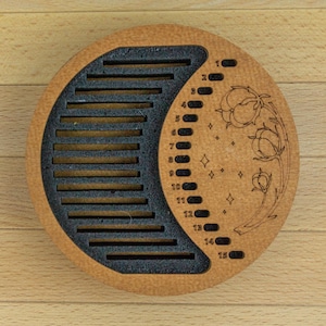 May include: A round wooden object with a crescent-shaped cutout and a series of horizontal slots. The other half features numbers 1-15, floral designs, and stars. The object is made of light brown wood with a dark gray crescent.