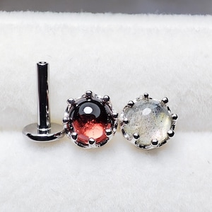 Pode incluir: Close-up de dois brincos de pressão com pedras preciosas. Um brinco apresenta uma pedra preciosa redonda, vermelha escura, enquanto o outro tem uma pedra clara e iridescente. Ambos são definidos em uma configuração prateada em forma de coroa.