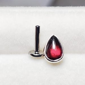 Op de afbeelding: Een zilveren traanvormig lichaamssieraad met een rode edelsteen. De sieraden liggen op een witte achtergrond.