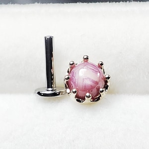 Puede incluir: Un labret de color plateado con una piedra veteada rosa y blanca, engastada en una corona dorada. La joya se exhibe sobre una superficie blanca.