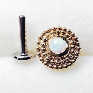 May include: A gold-coloured lip piercing stud featuring a central opal stone, encircled by small, beaded details. Beside it is a silver-coloured post with a flat, circular base. The jewellery is presented on a white, textured surface.