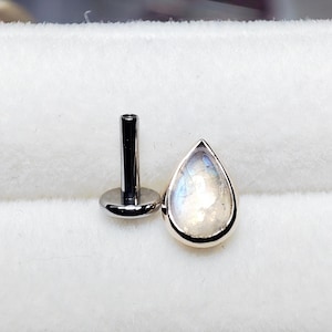 May include: A teardrop-shaped, iridescent moonstone stud earring with a silver bezel and post. The moonstone displays a play of color, with hints of blue and white. The earring is set against a white background.